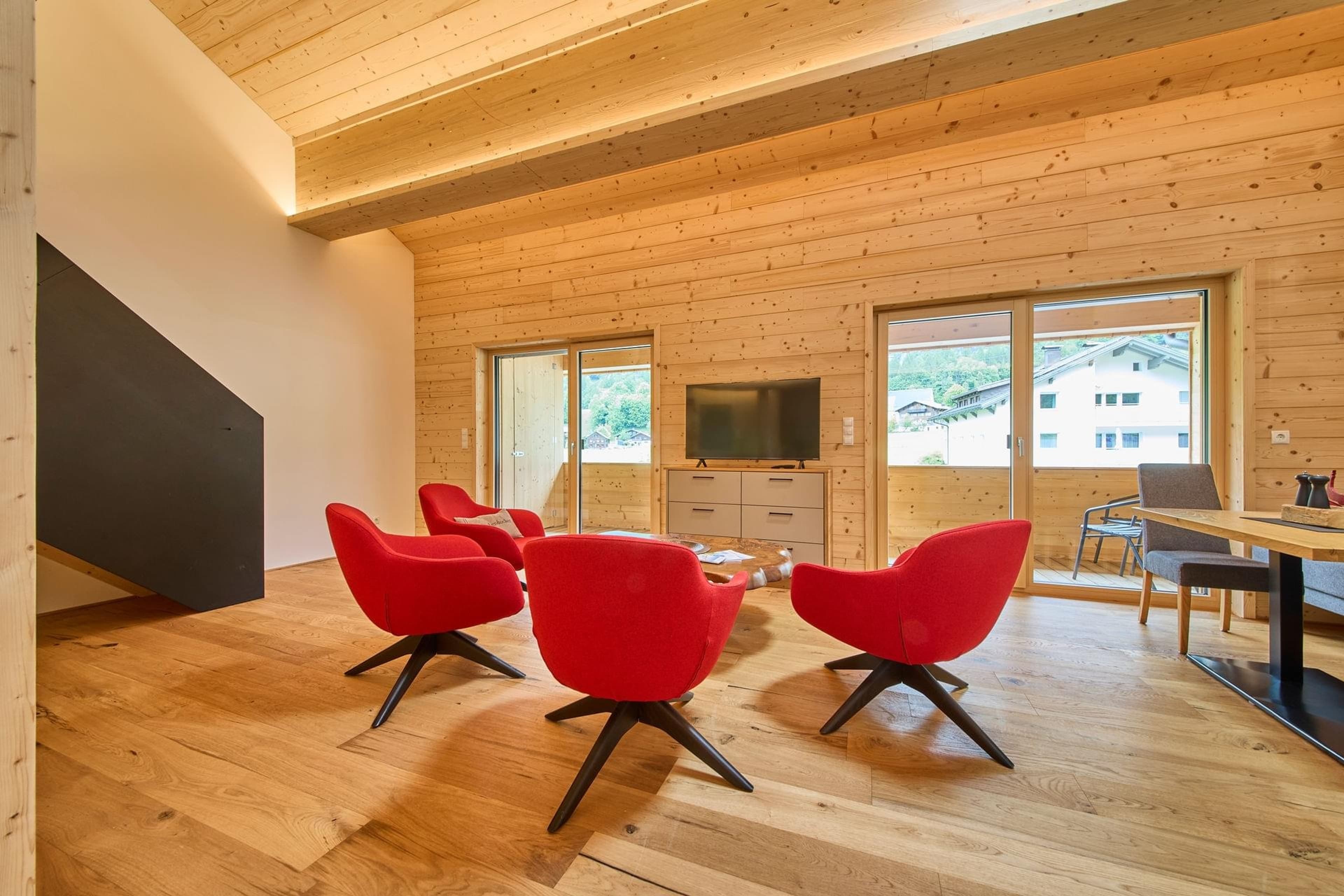 Bocks Apartments 53C - Top 4 Fichte - Bright and spacious living area with a TV, a dining table with chairs, and a seating area with vibrant red armchairs. A large wooden coffee table is a central feature. Large windows offer views to the outside, and wooden floors complement the overall design. The atmosphere is welcoming and contemporary.